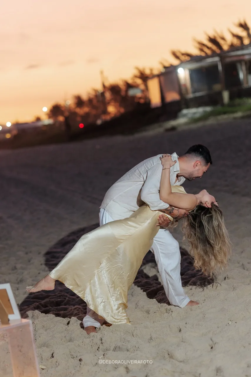 Eduardo inclinando Isabely para um beijo na praia ao pôr do sol